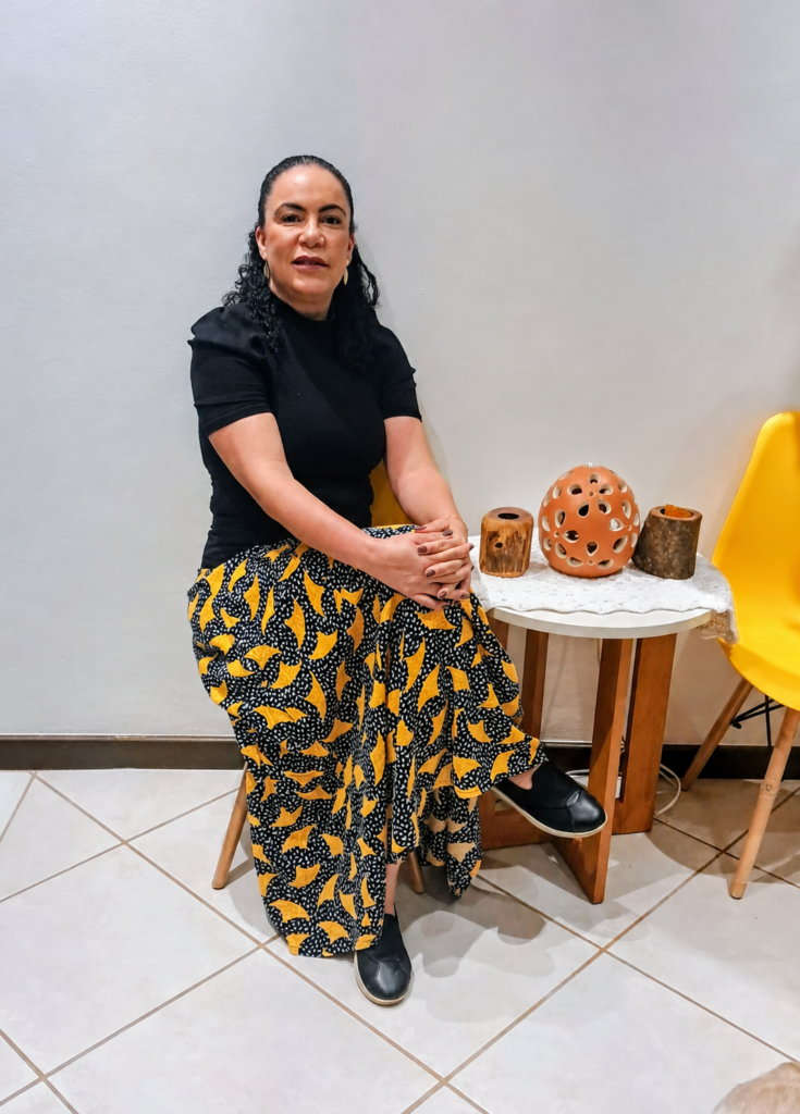 Dra. Denise Lopes sentada, com roupa preta e saia estampada em amarelo, em retrato de corpo inteiro