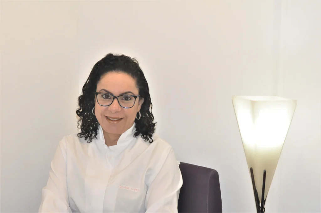 Dra. Denise Lopes de jaleco branco e óculos, em retrato no ambiente de atendimento